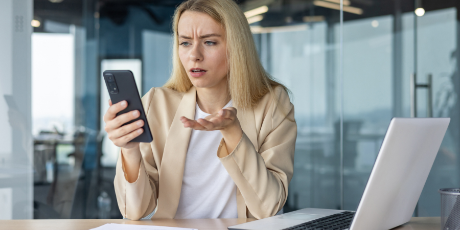 Worried young business woman working in the office at the laptop, holding the phone in her hands, anxiously looking at the screen - stock photo